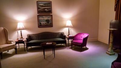 Dim living room with a dark sofa, two lamps, framed art, and a bright pink armchair.