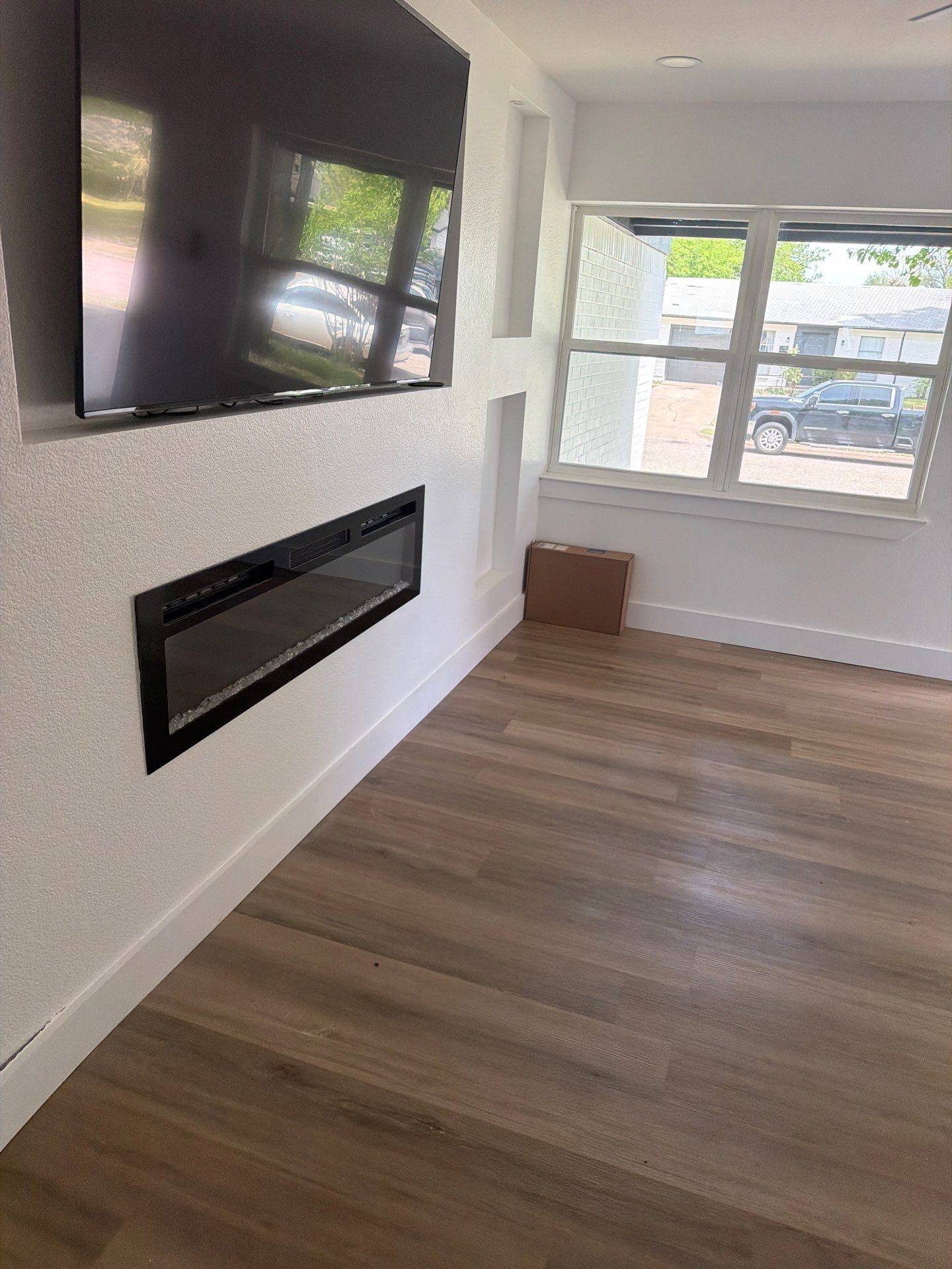 Empty modern living room with wall-mounted TV, fireplace, wood floors, and a large window.
