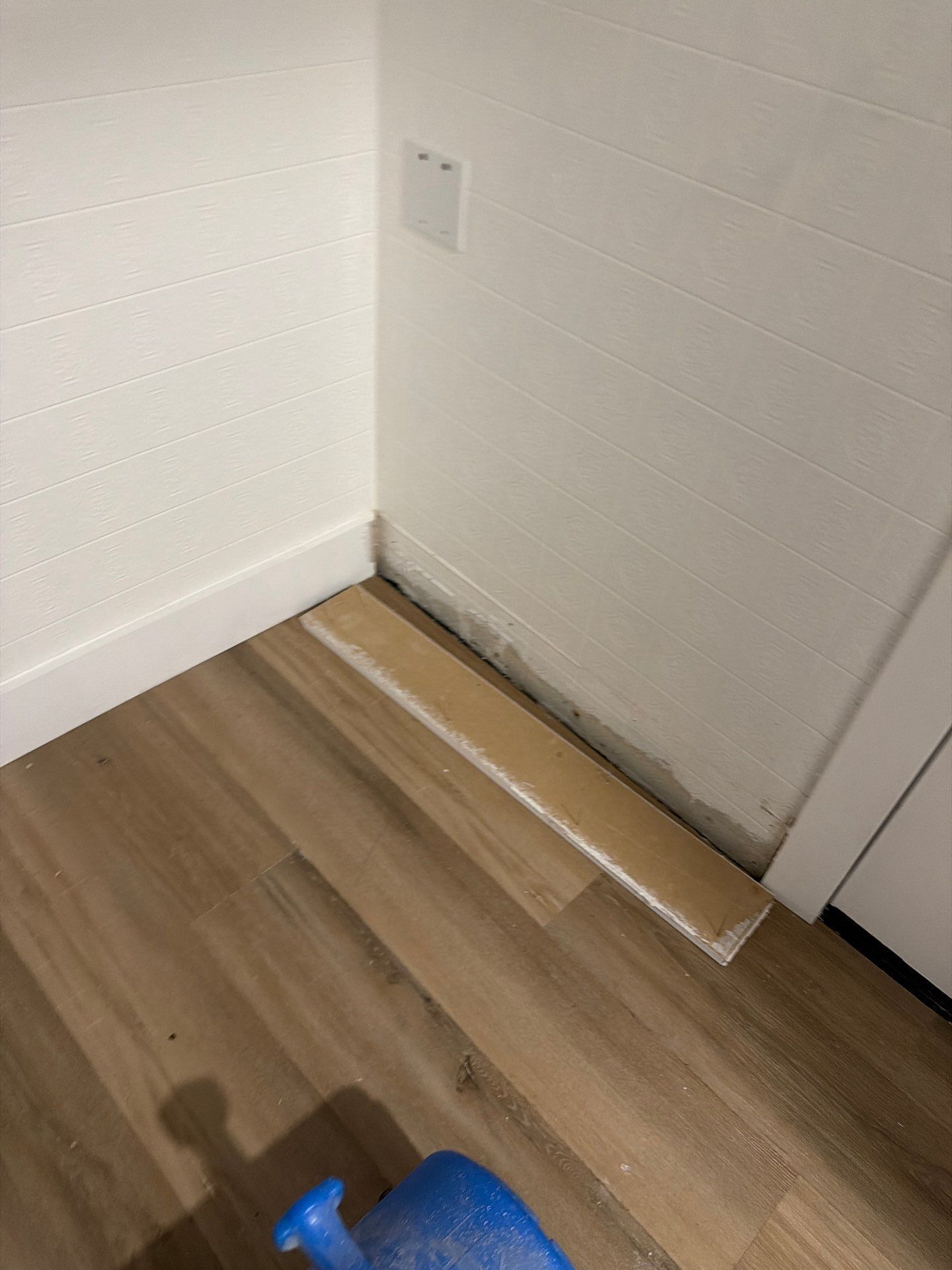 Corner of a room with a wooden floor, white walls, and a white door; a blue spray bottle sits at the bottom.