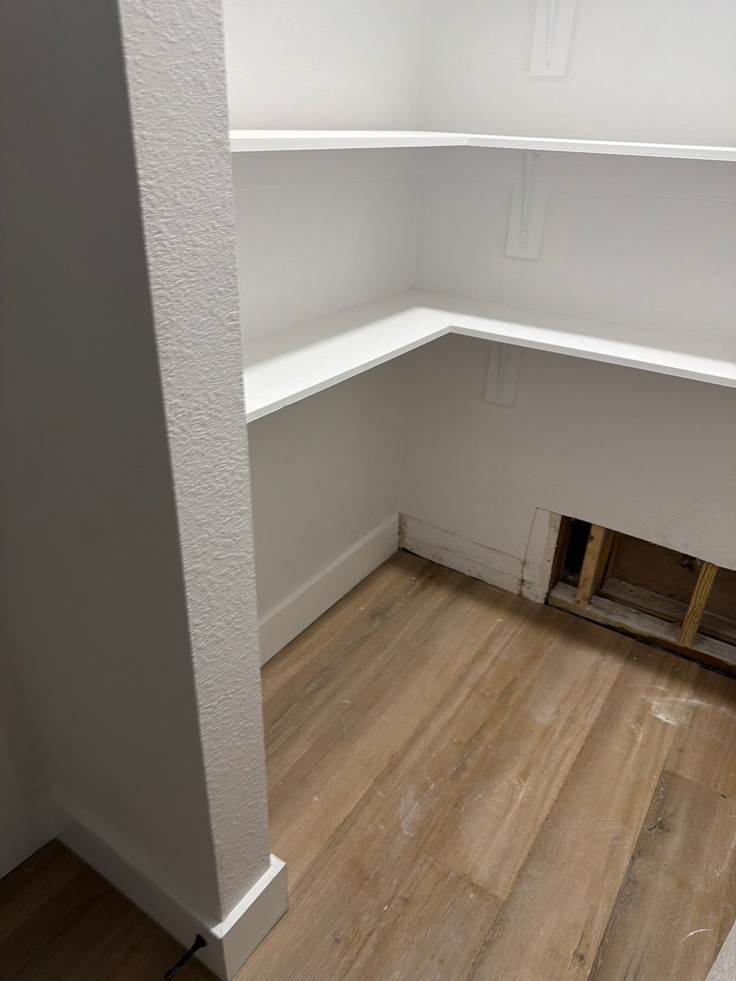 Empty walk-in closet with white shelves and wood-look flooring; open wall section exposed on right.