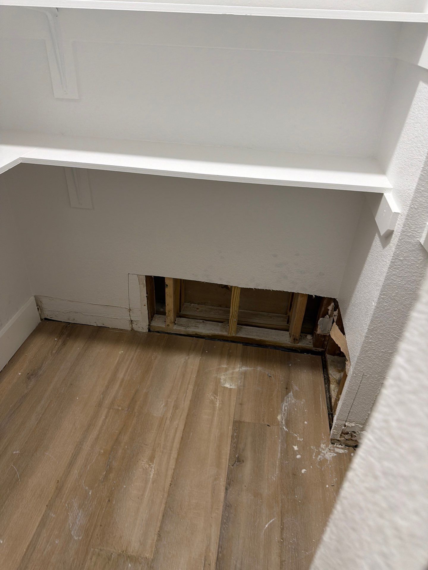 Damaged wall opening in a room with wood flooring and white trim, exposing studs and wiring