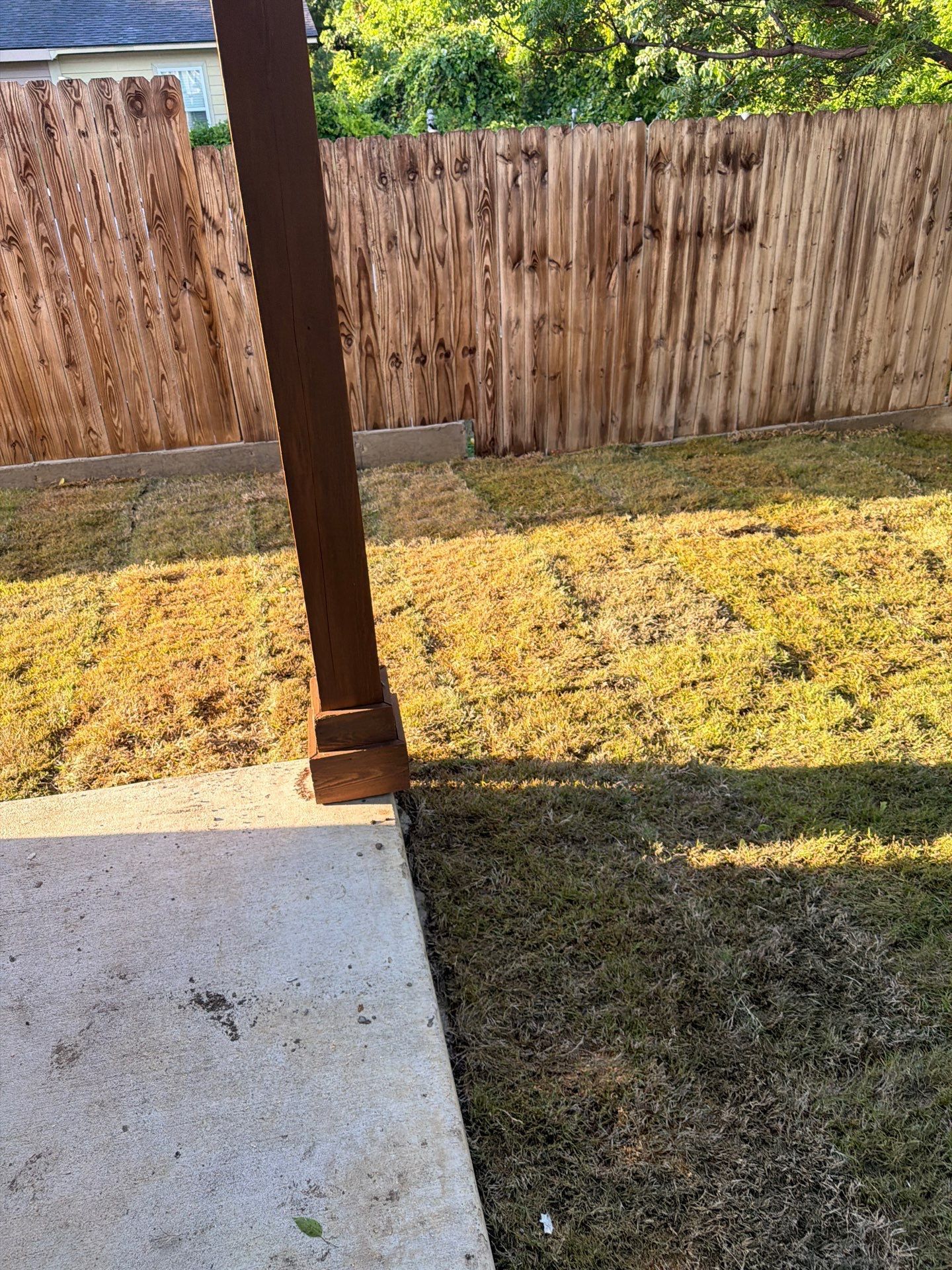 Backyard patio with a wooden fence, grassy yard, and a brown post casting a shadow.