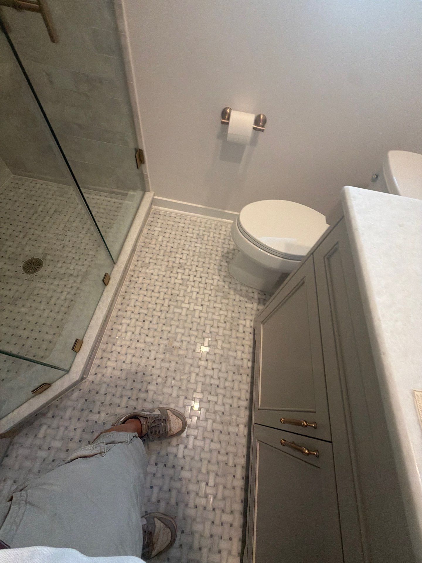 Small bathroom with glass shower, white tile floor, toilet, vanity, and a person’s feet visible.