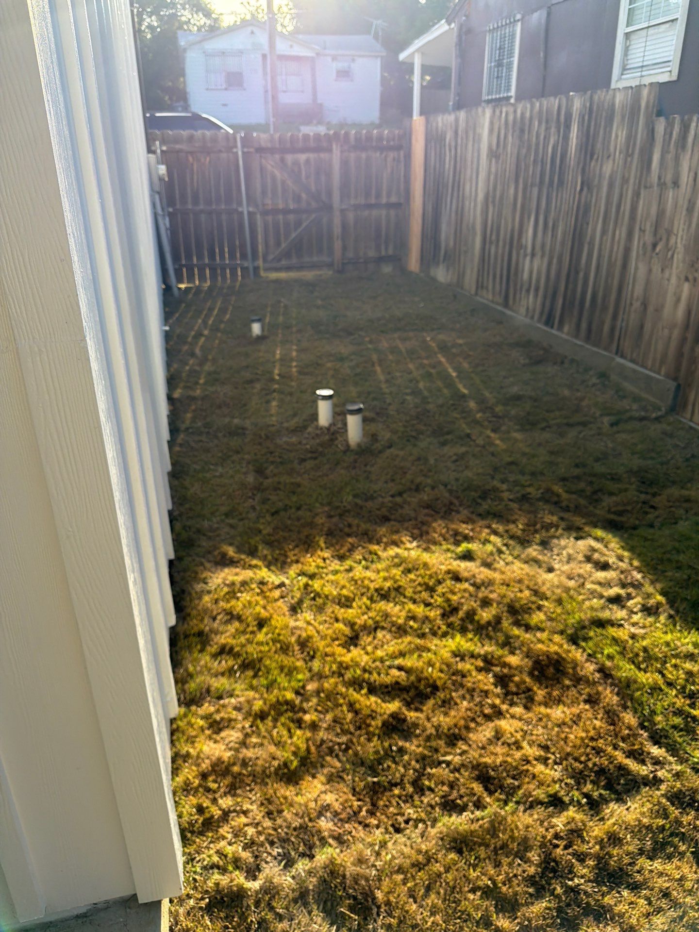Narrow fenced backyard with patchy grass, three white pipes, and sunlight near the house wall