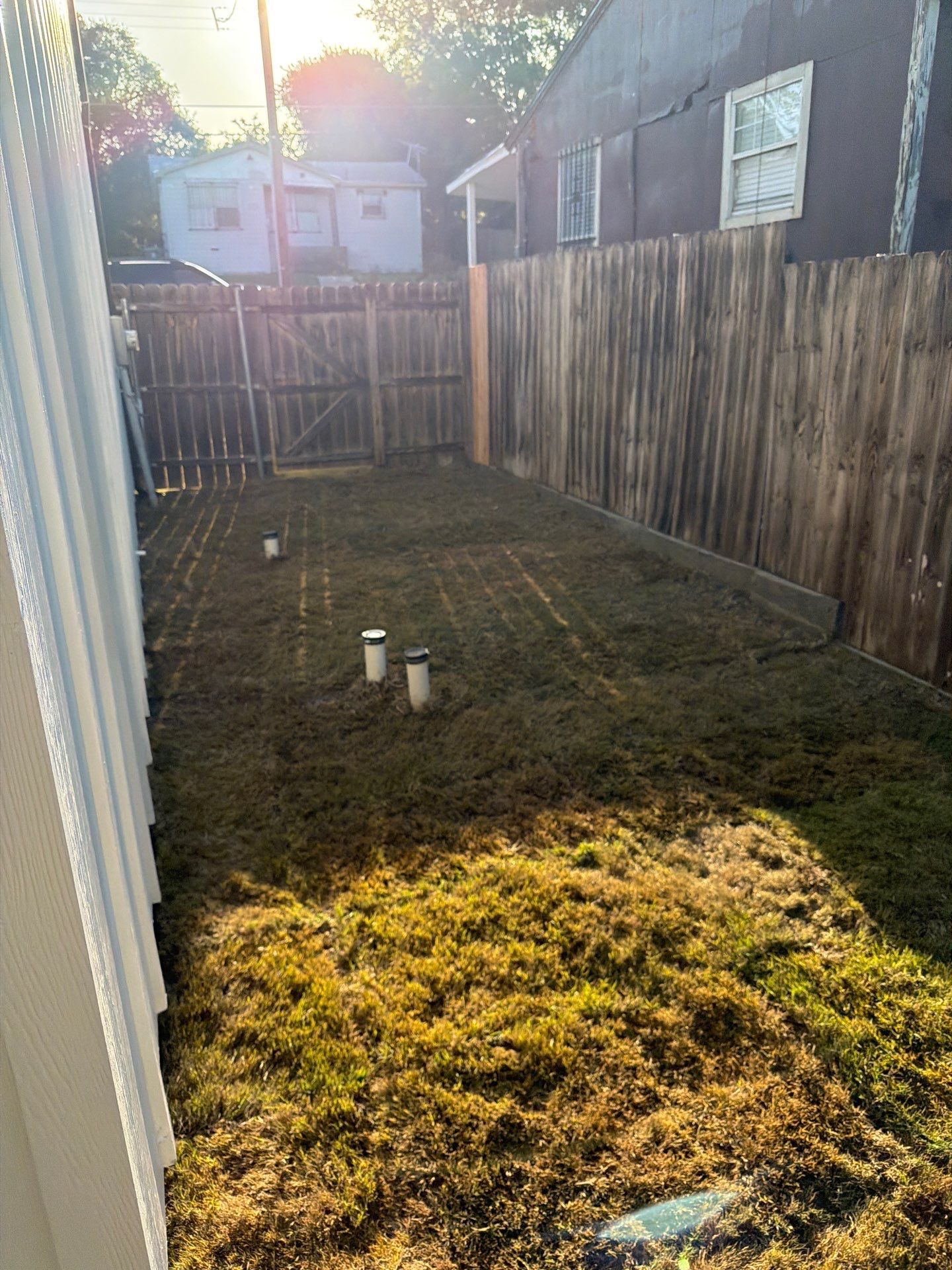 Narrow fenced backyard with patchy grass, a small concrete path, and neighboring houses in the distance
