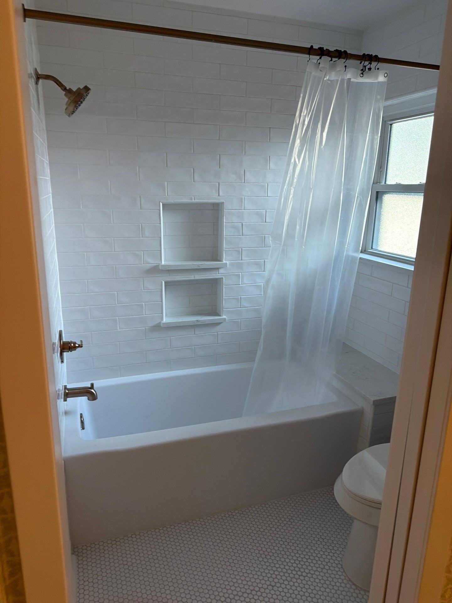 Small bathroom with white tiled tub, shower curtain, window, and toilet.
