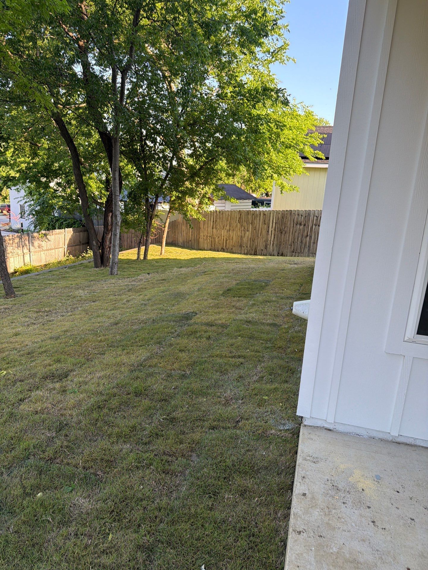 Backyard with grass, trees, wooden fence, and white house siding on the right