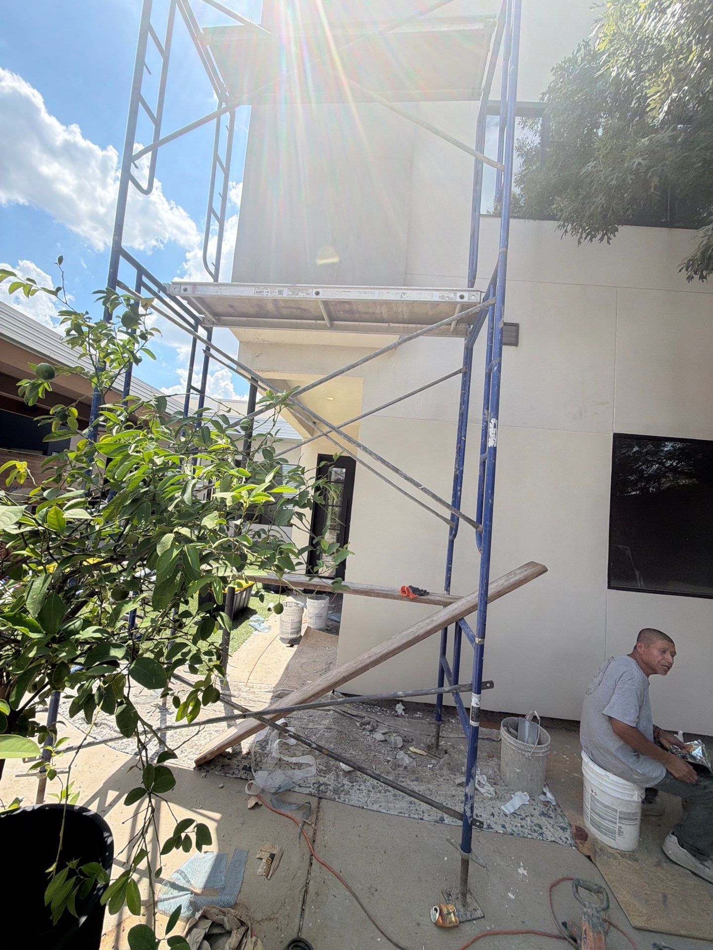 Scaffolding beside a white building, with a worker mixing material on the ground in bright sunlight.