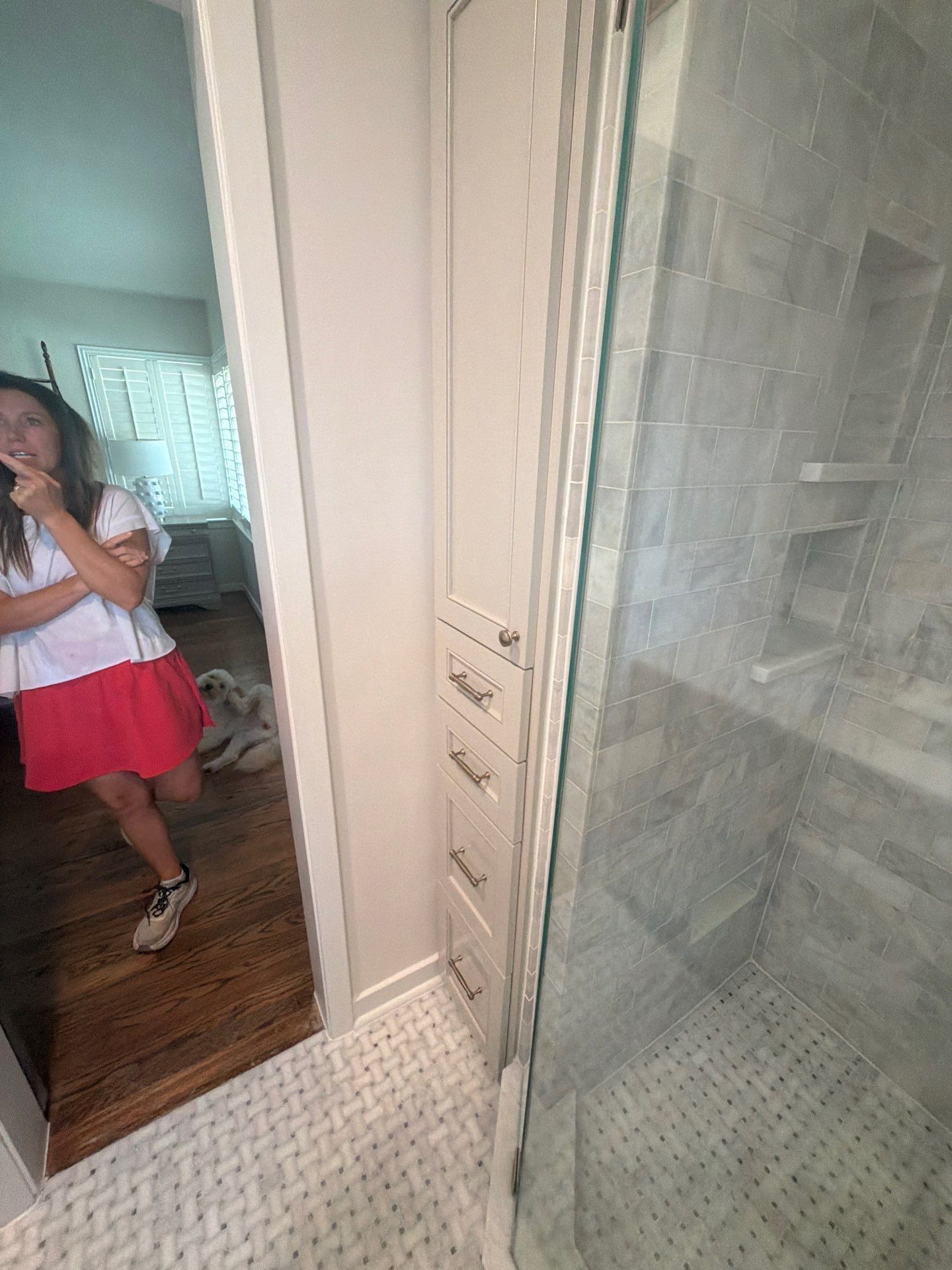 Bathroom doorway with a mirror, shower, and a person in a white top and pink skirt reflected in the hall mirror