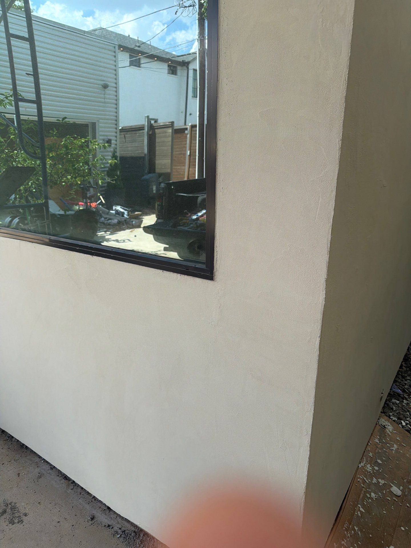 Exterior wall with a window reflecting a backyard and neighboring building, with a blurred finger in the foreground