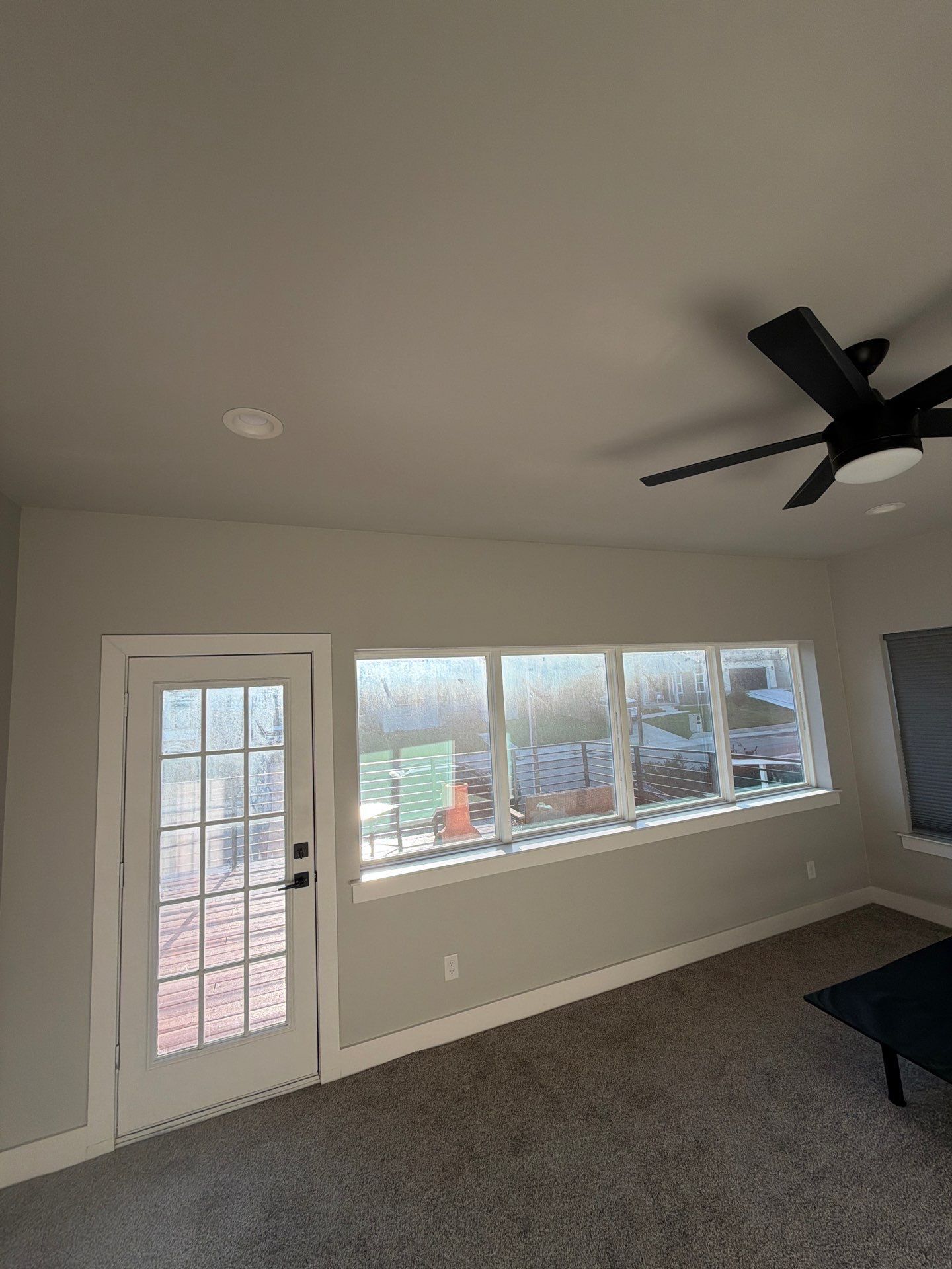 Empty beige living room with carpet, ceiling fan, and large windows beside a glass door