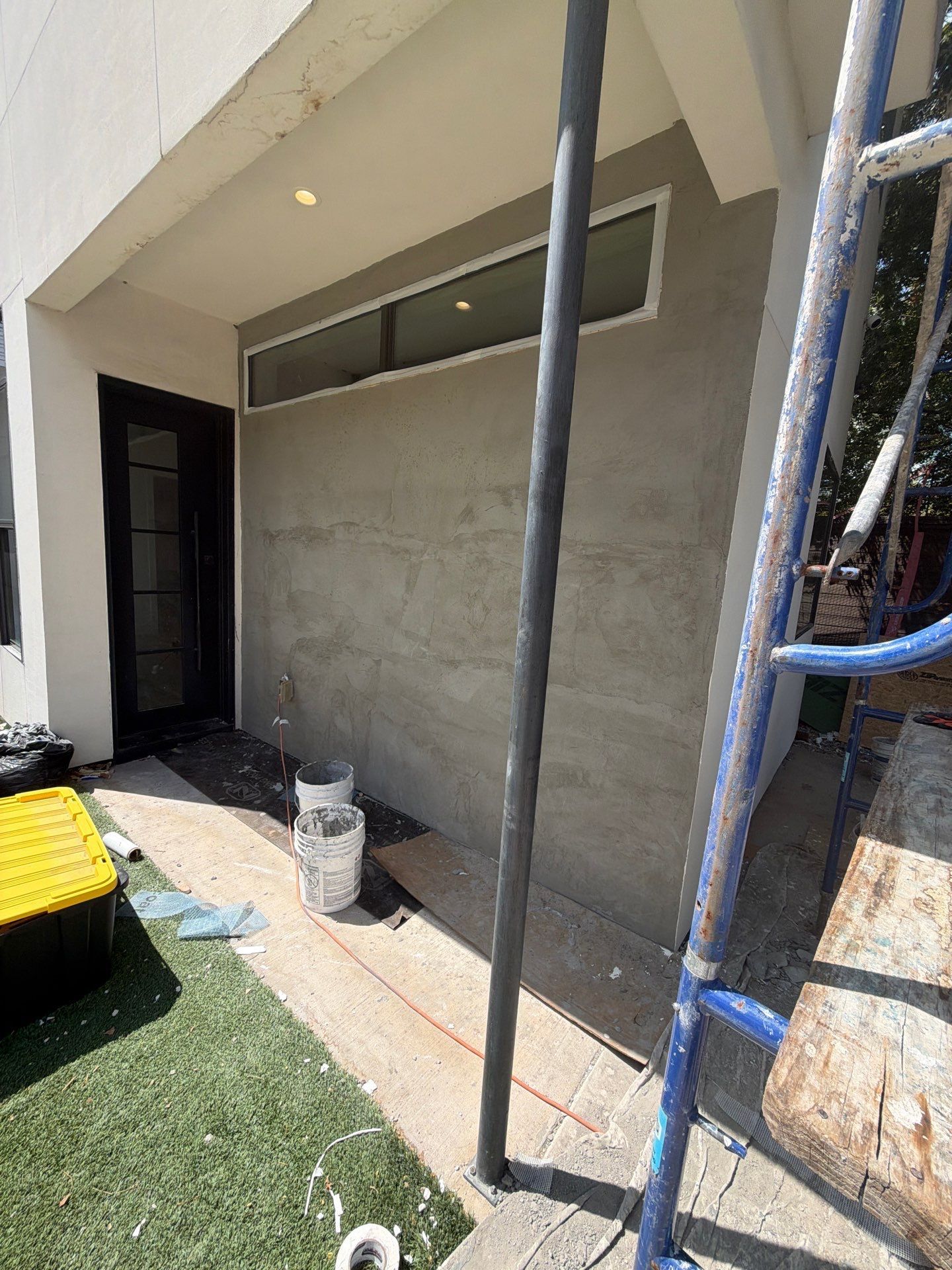 Gray stucco wall under construction with scaffolding, ladder, and a doorway beside artificial turf.