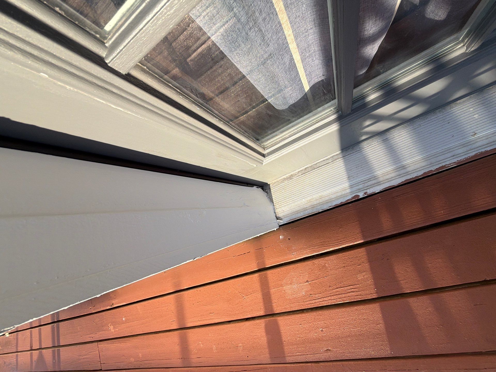 Close-up of a house corner with white trim, a glass window, and red siding in sunlight.