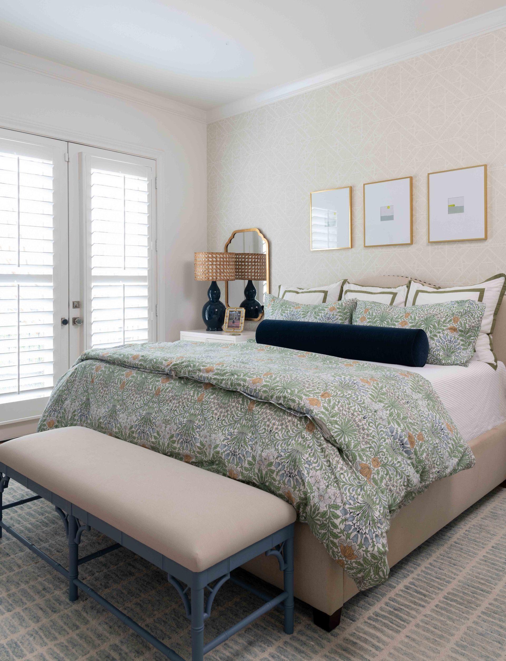 Bright bedroom with a bed, bench, patterned bedding, framed wall art, and white shutters by the window