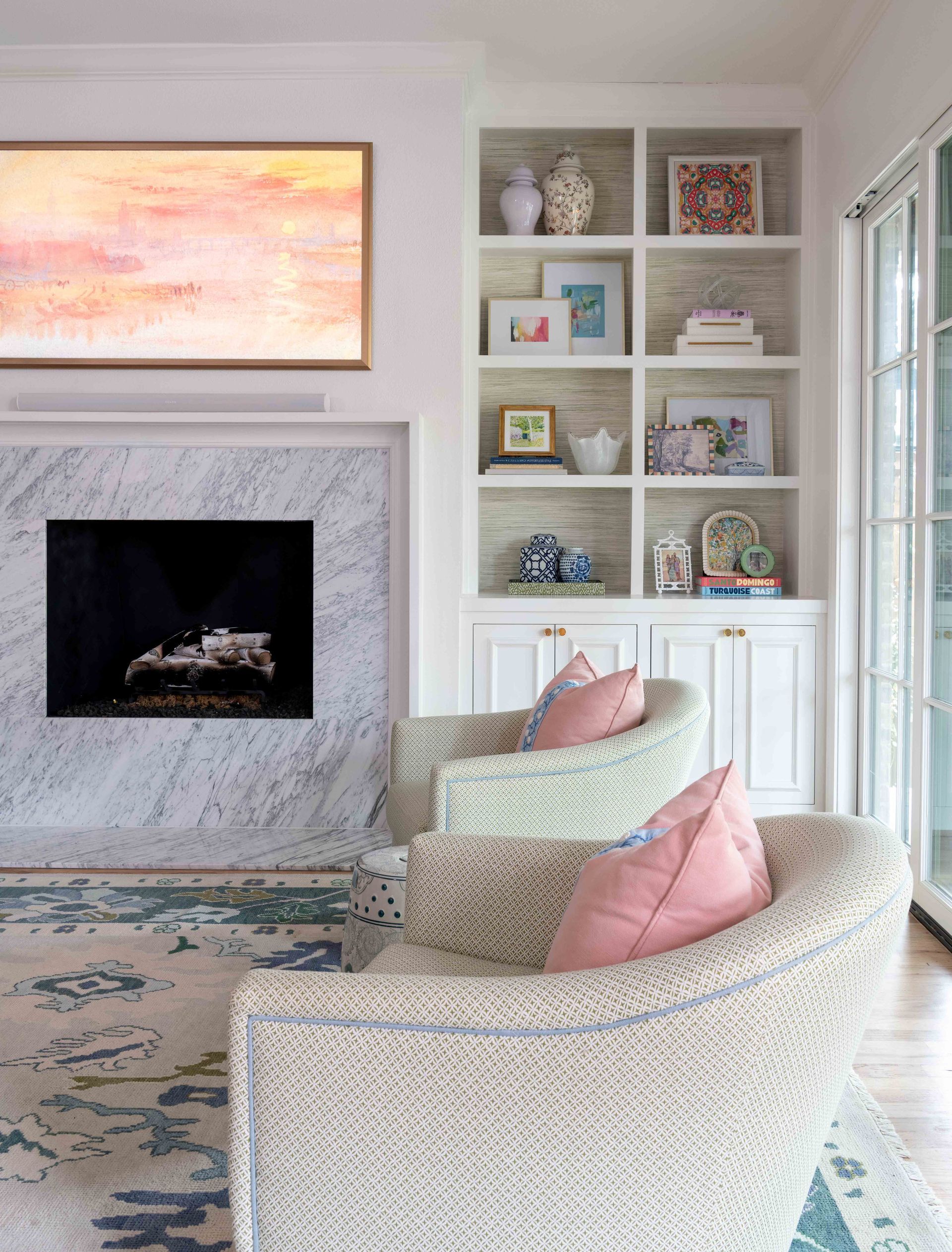 Cozy living room with woven chairs, pink pillows, marble fireplace, and built-in shelves with decor.