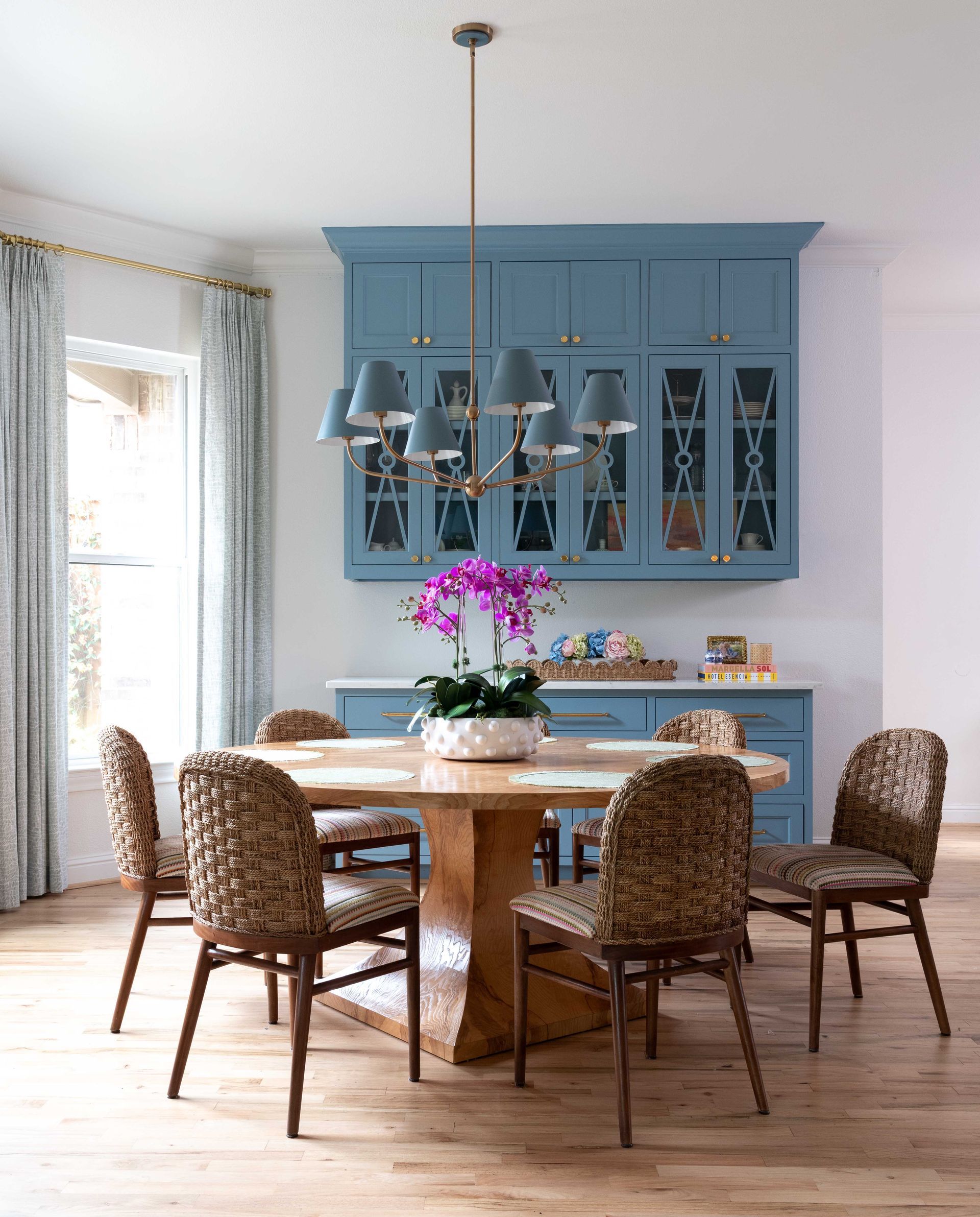 Blue dining room with wooden table, woven chairs, hanging pendant lights, and a flower centerpiece