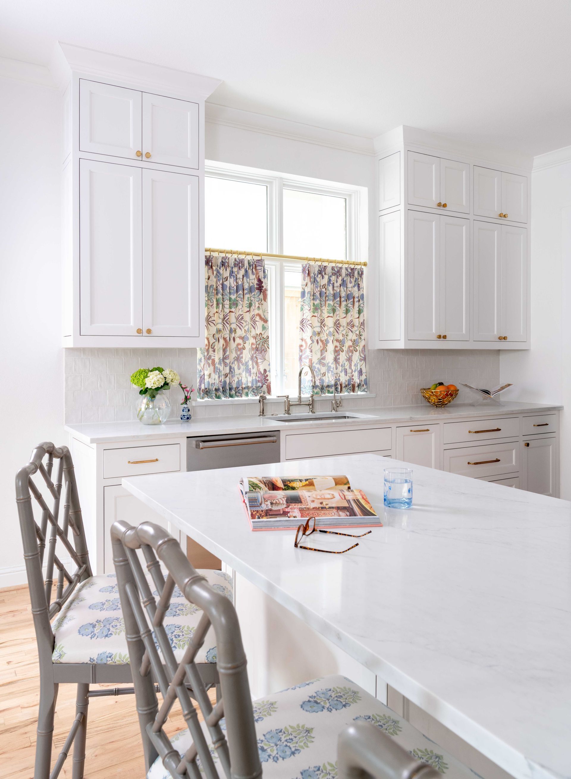 Bright white kitchen with an island, two chairs, and a window over the sink with floral curtains.