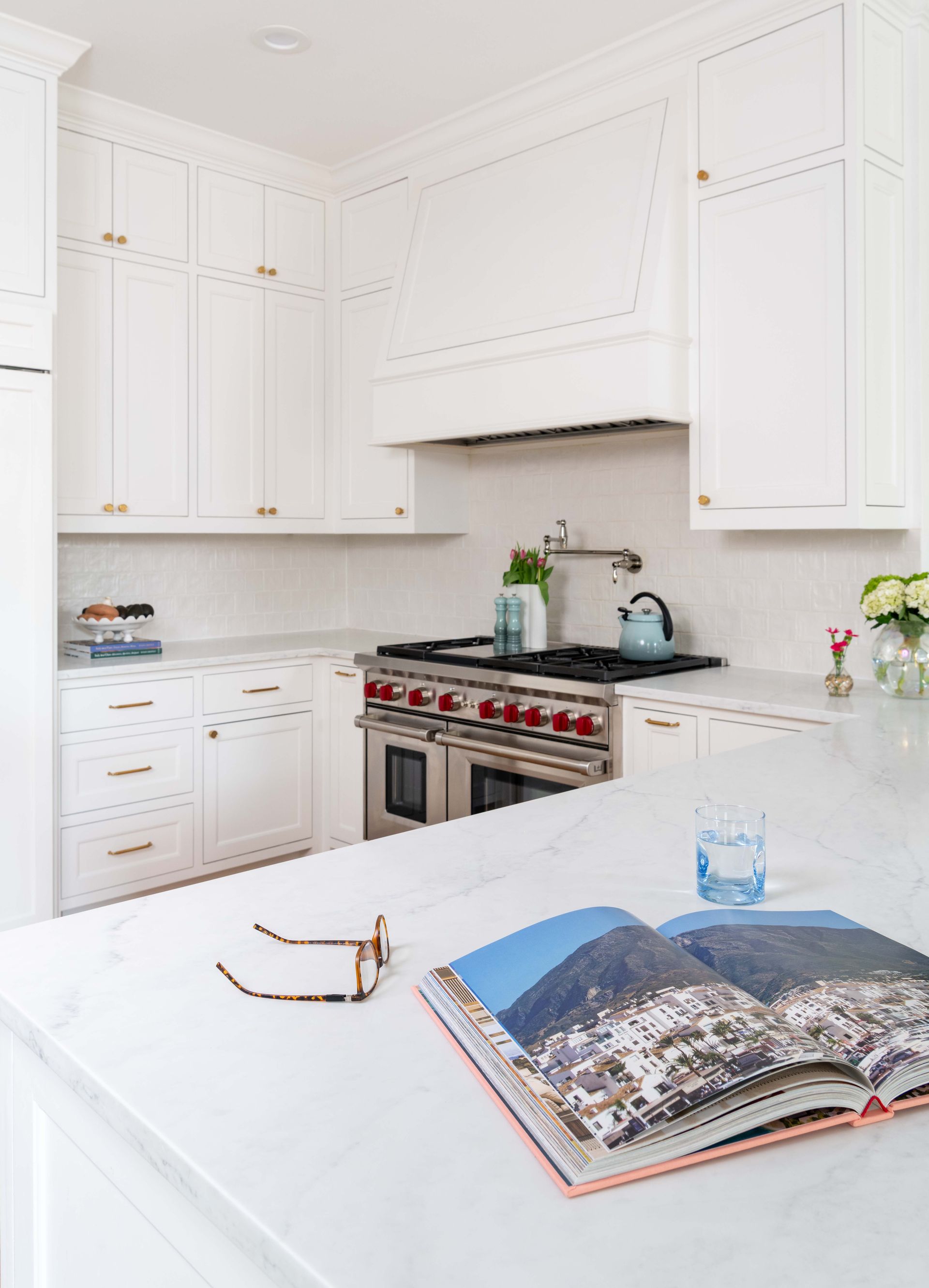 Bright white kitchen with stainless steel stove, open cookbook, and reading glasses on a countertop