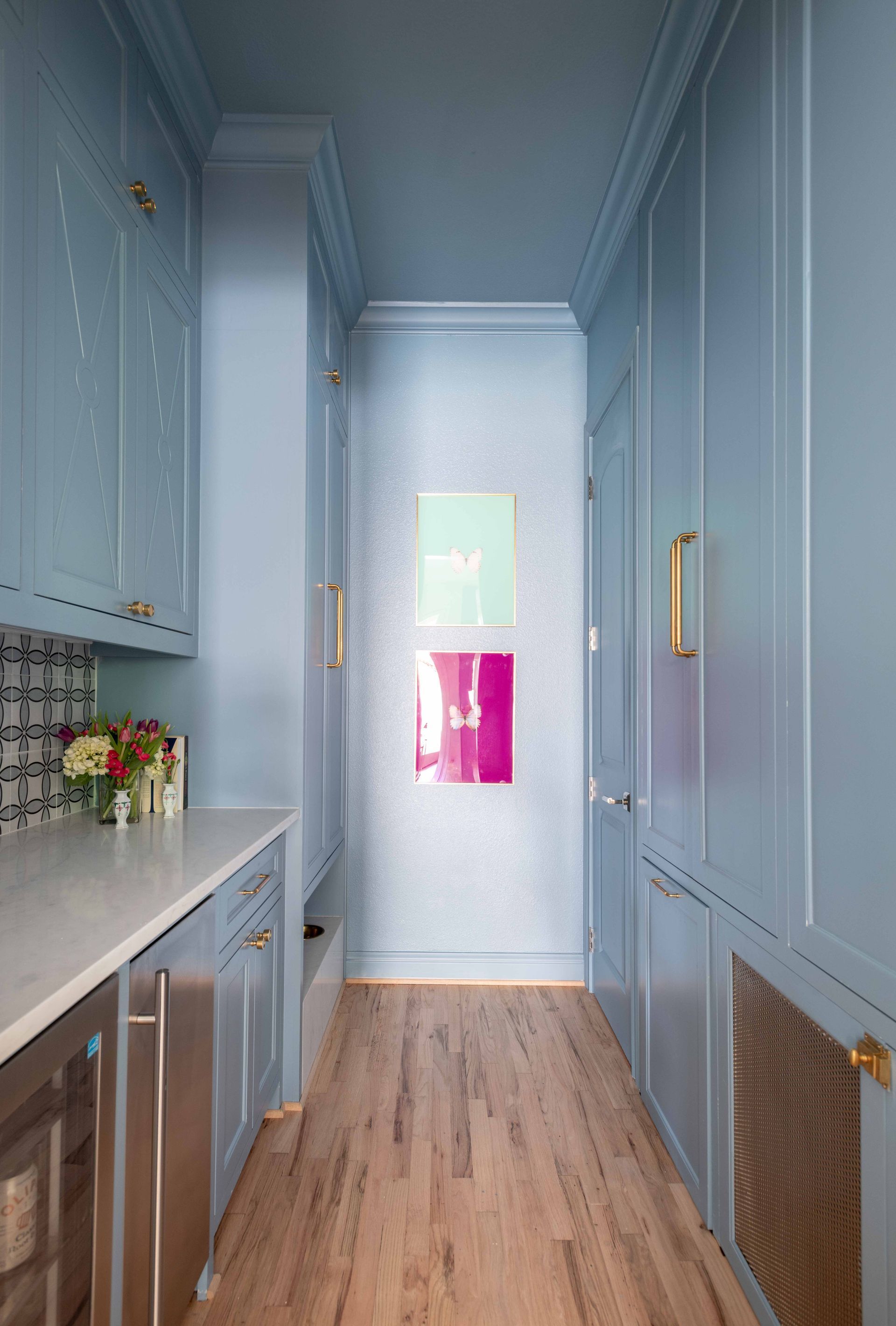 Narrow pastel-blue galley kitchen with white counters, wood floor, and a bright window at the end