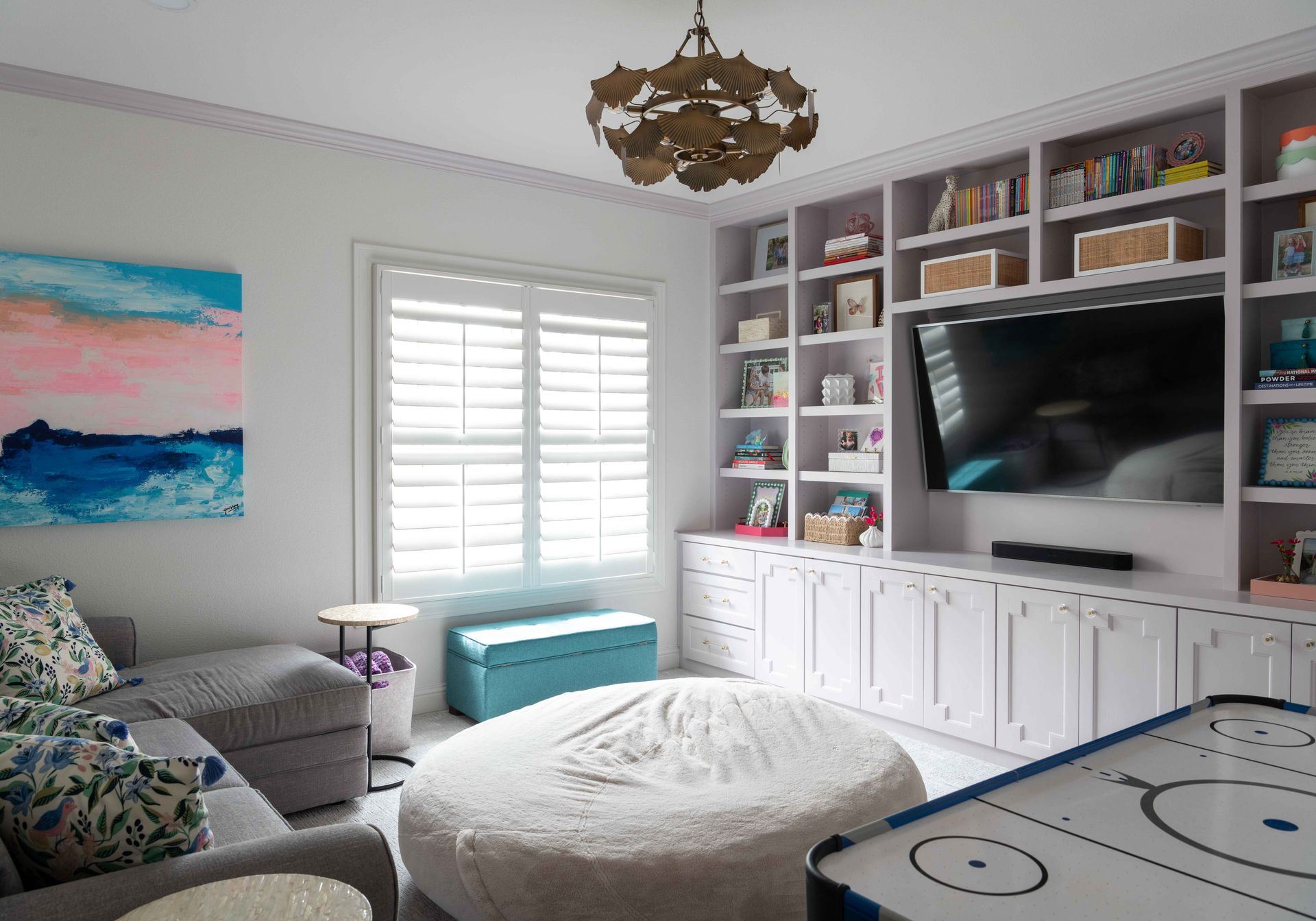 Bright playroom with white built-ins, TV, window seat, teal ottoman, round pouf, and colorful wall art.
