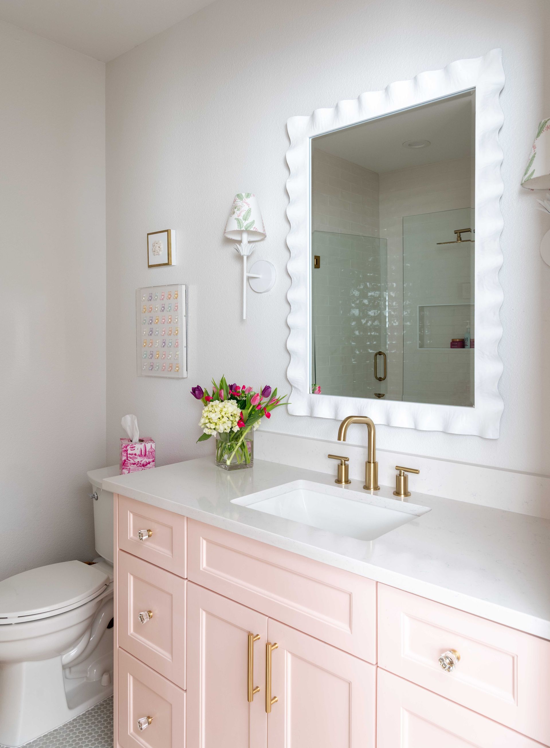 Soft pink bathroom vanity with gold fixtures, white countertop, mirror, and flowers beside a toilet.