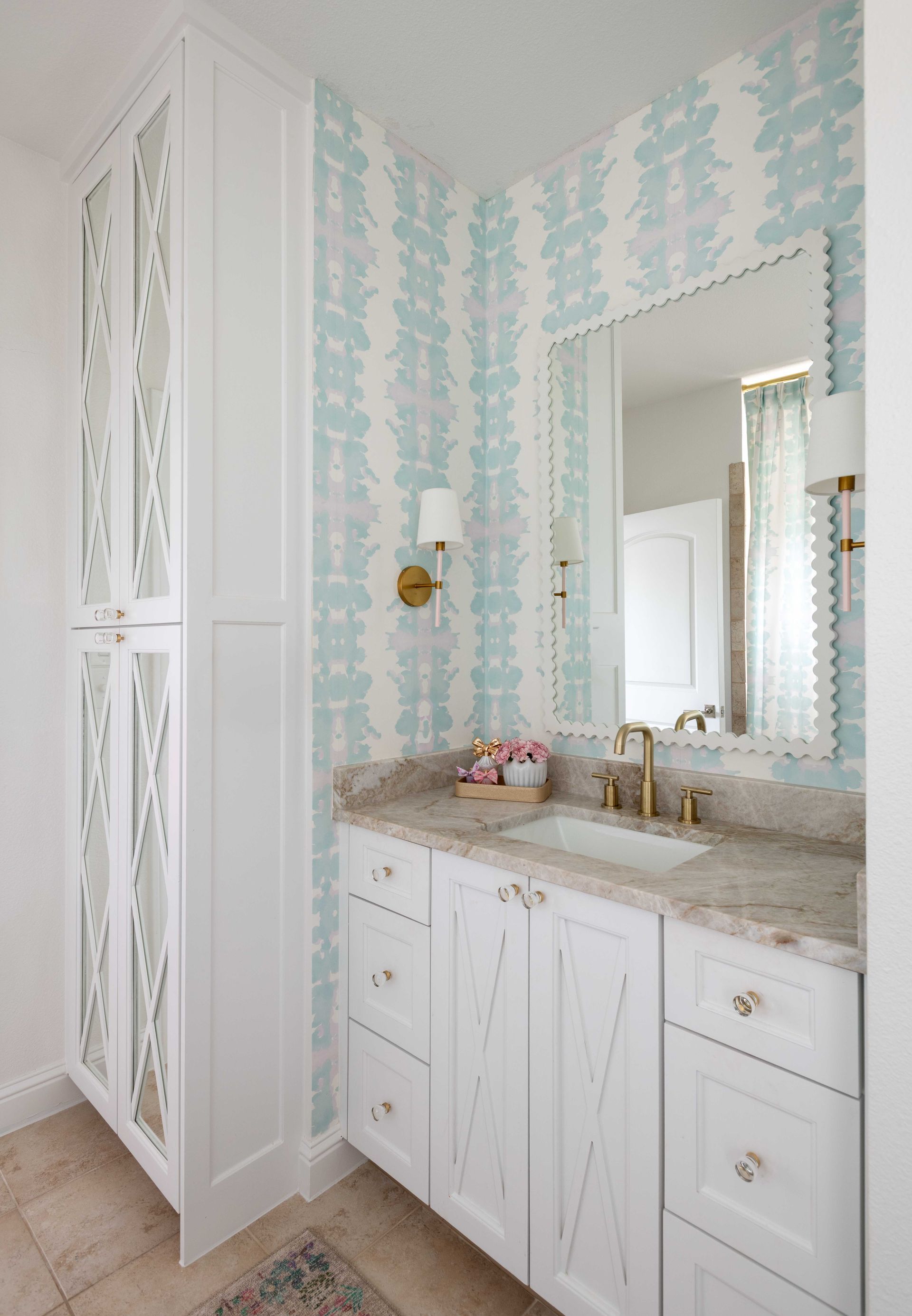 Bright bathroom vanity with marble countertop, white cabinets, gold fixtures, and blue patterned wallpaper
