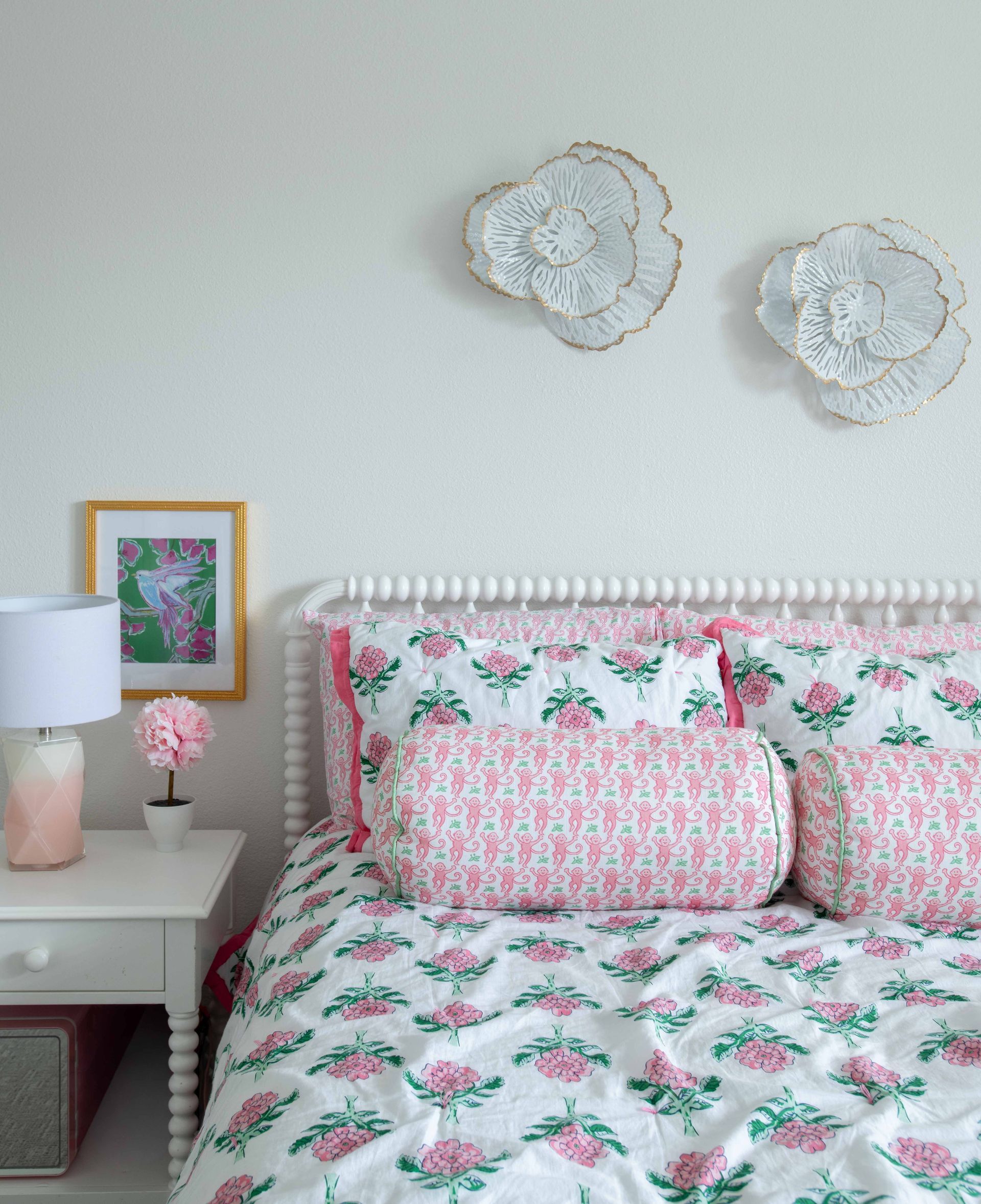 Pink floral bedspread in a pastel bedroom with white walls, wall art, and a bedside table lamp.