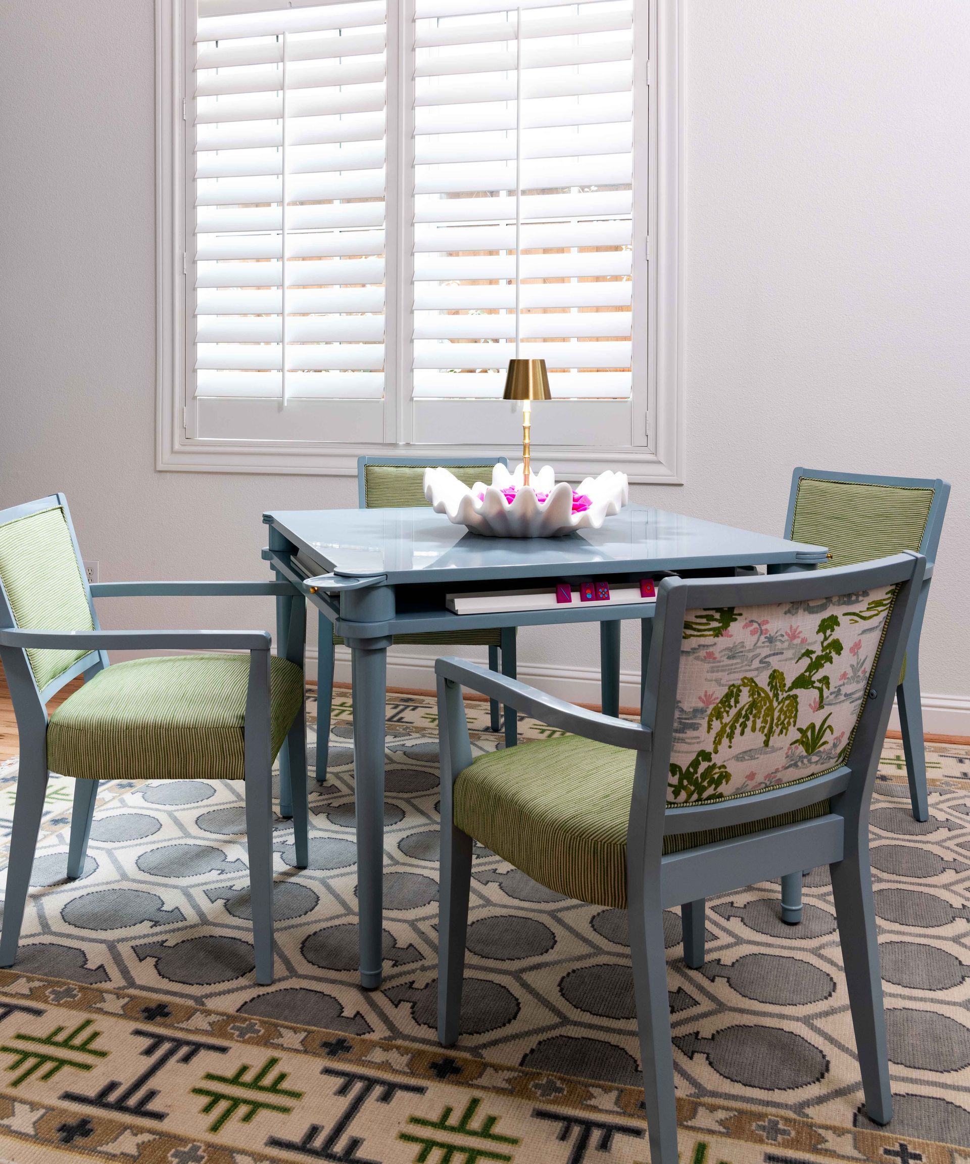 Patio table with four gray chairs and patterned cushions in a bright corner by shuttered windows.