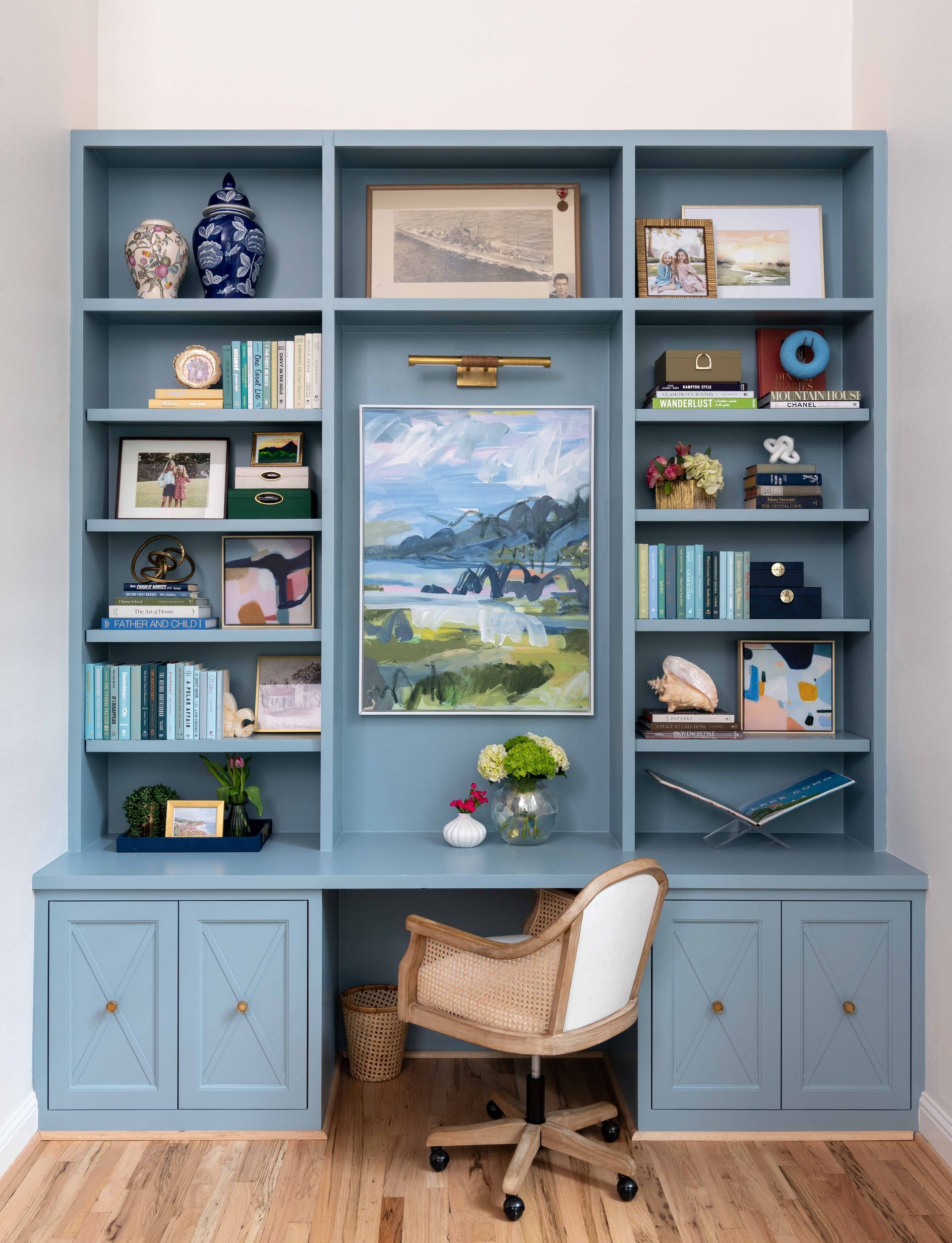 Blue built-in shelves with books, decor, and a central landscape painting above a wicker chair and cabinet drawers.