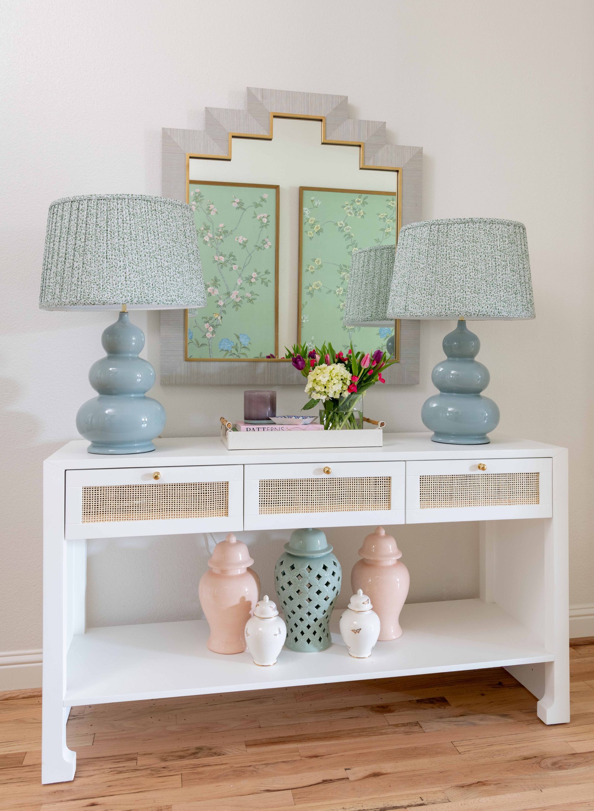 White console table with pastel vases, lamps, flowers, and a gold-framed mirror against a light wall.