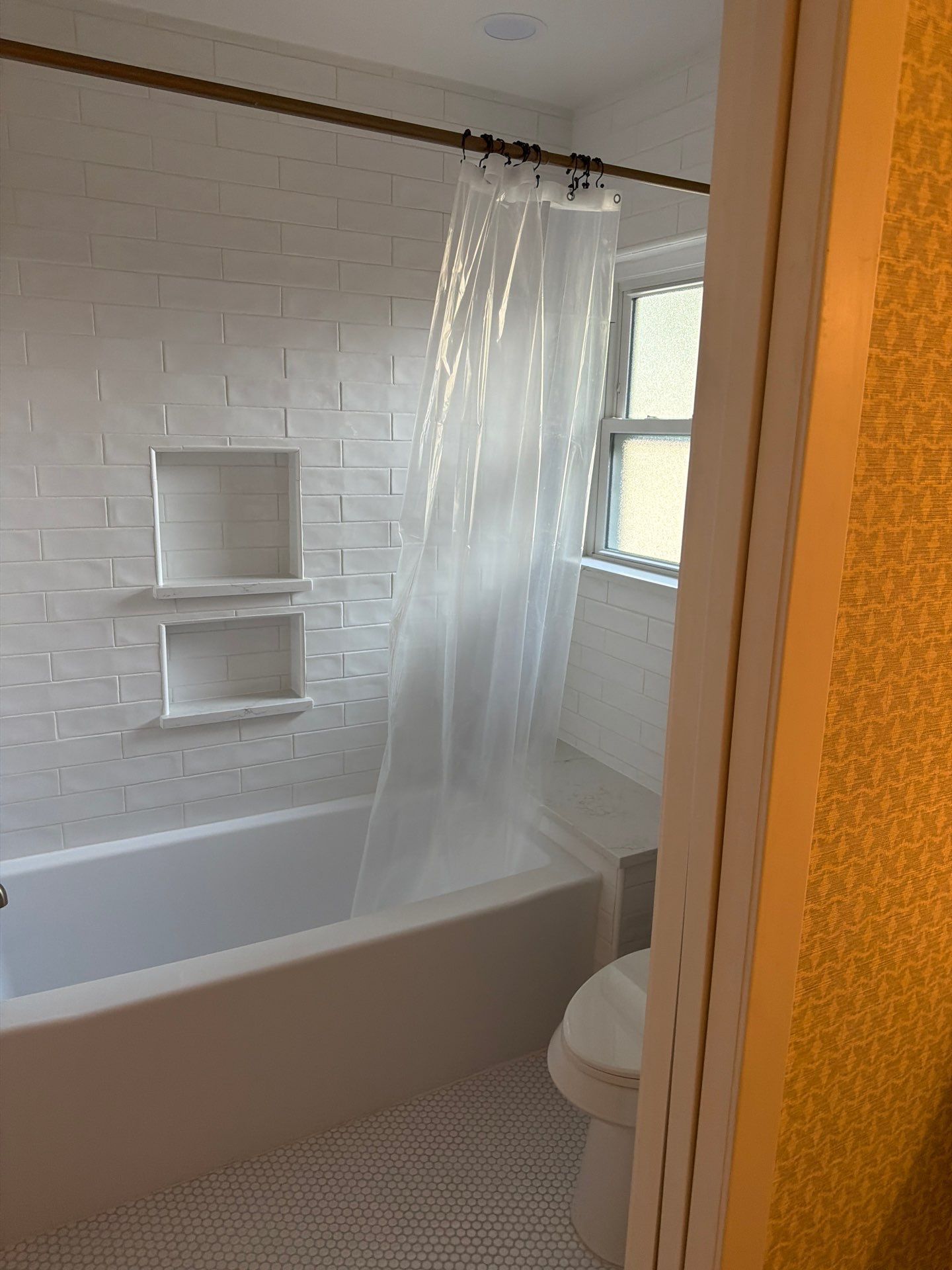Small bathroom with a white bathtub, shower curtain, wall shelves, and a toilet by the window.