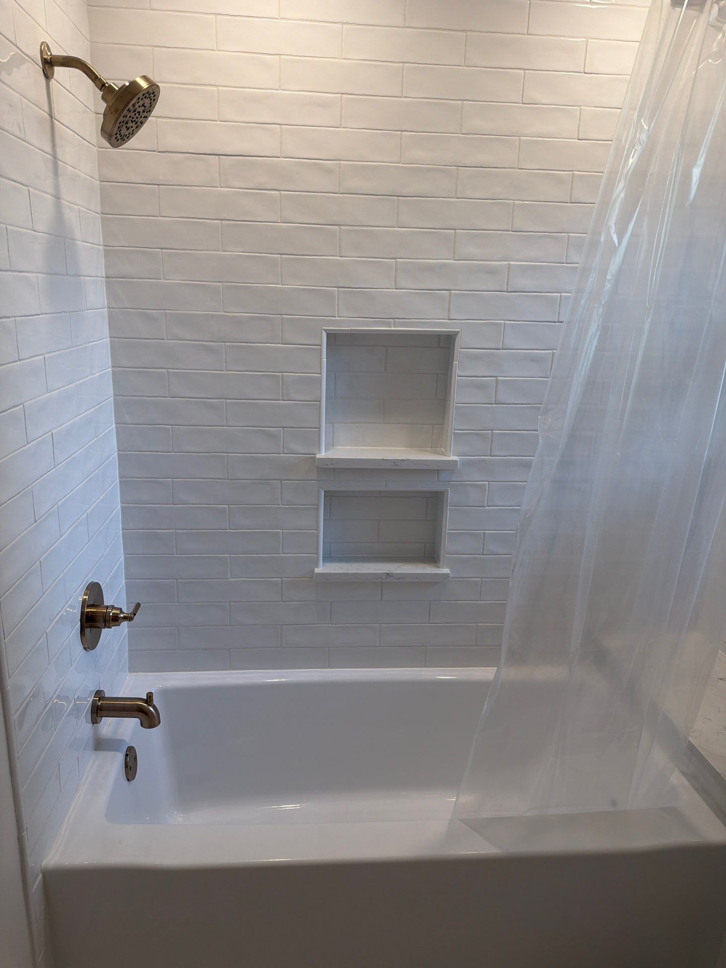 Small white tiled bathtub and shower with built-in wall niches and clear curtain.