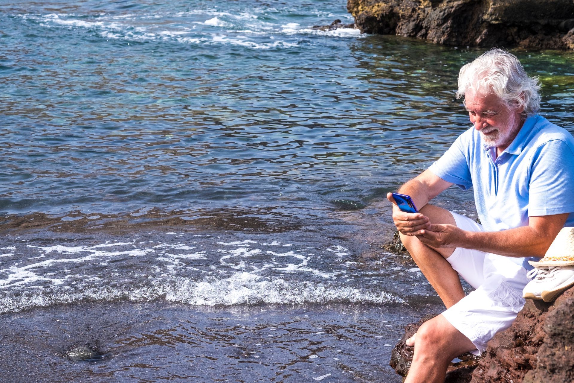 An older man is sitting on a rock near the water looking at his cell phone.