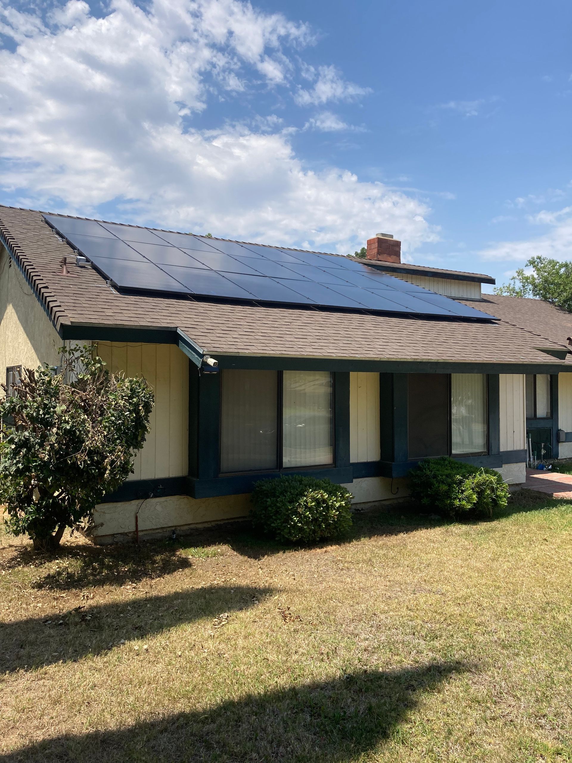 Solar panels on a residential roof against a blue sky, beige house with shrubs and brown trim.
