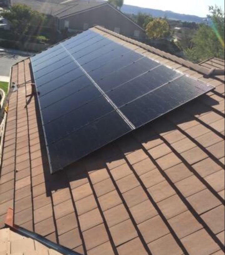 Solar panels installed on a brown-tiled roof of a house. Sunny day with a residential street visible.