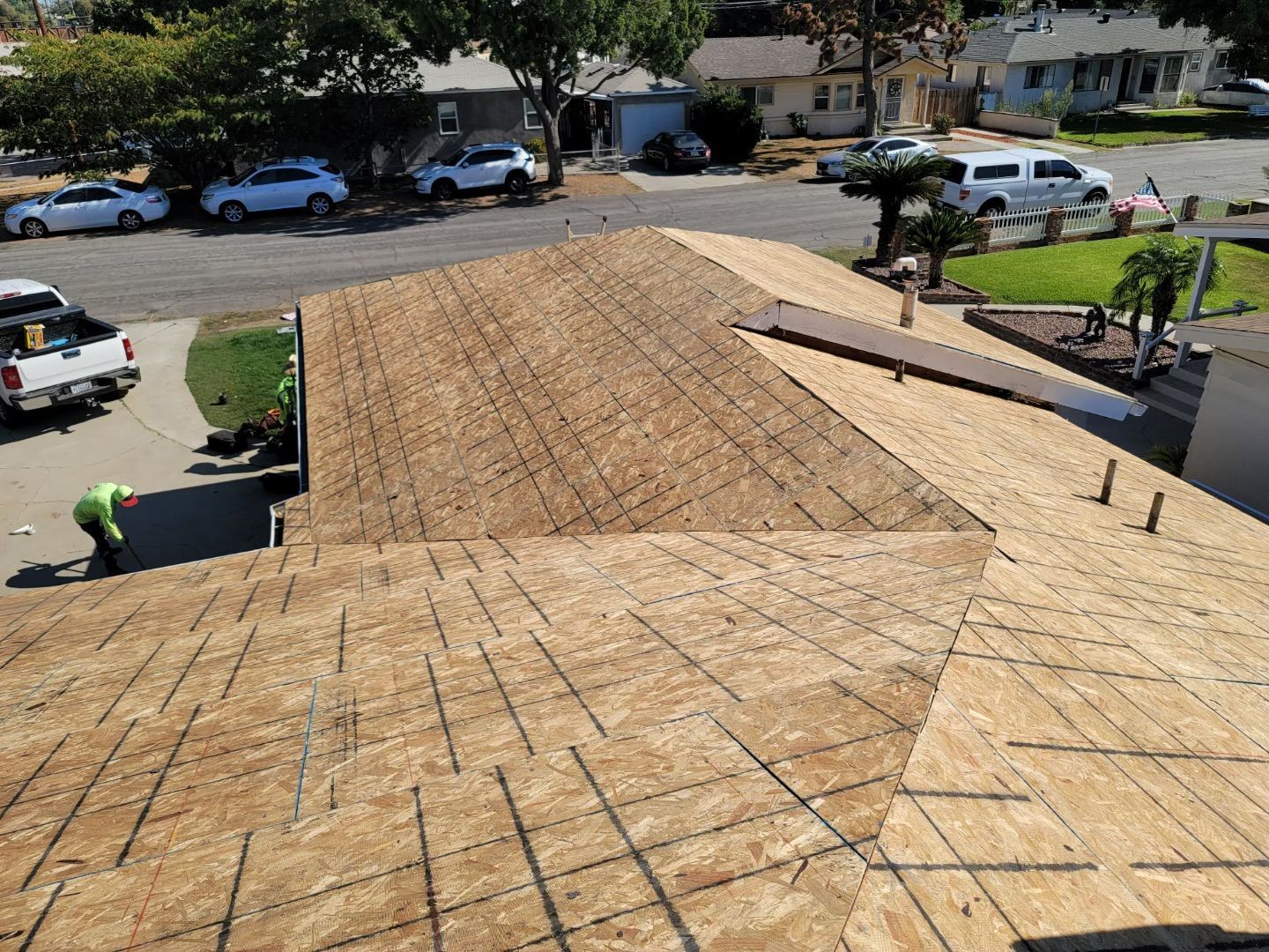 Roof being re-shingled, with exposed plywood and workers in a suburban neighborhood.