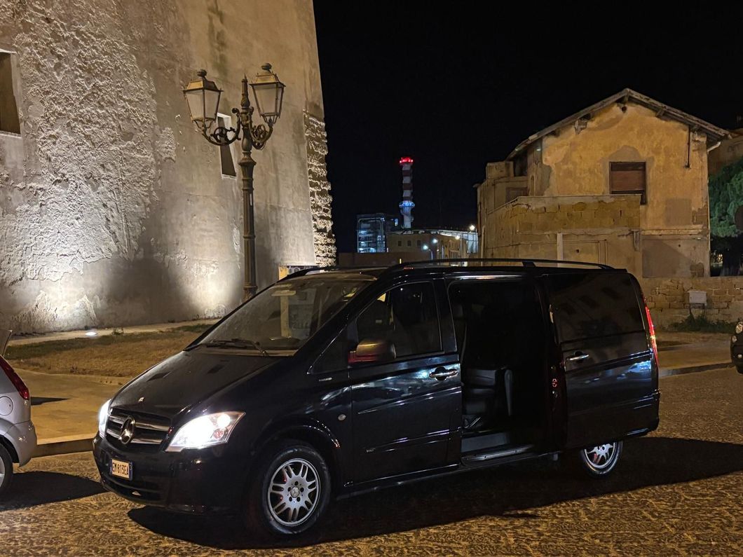 Un furgone nero parcheggiato al crepuscolo davanti a una villa in pietra illuminata da una luce calda, con archi ricoperti di fiori.