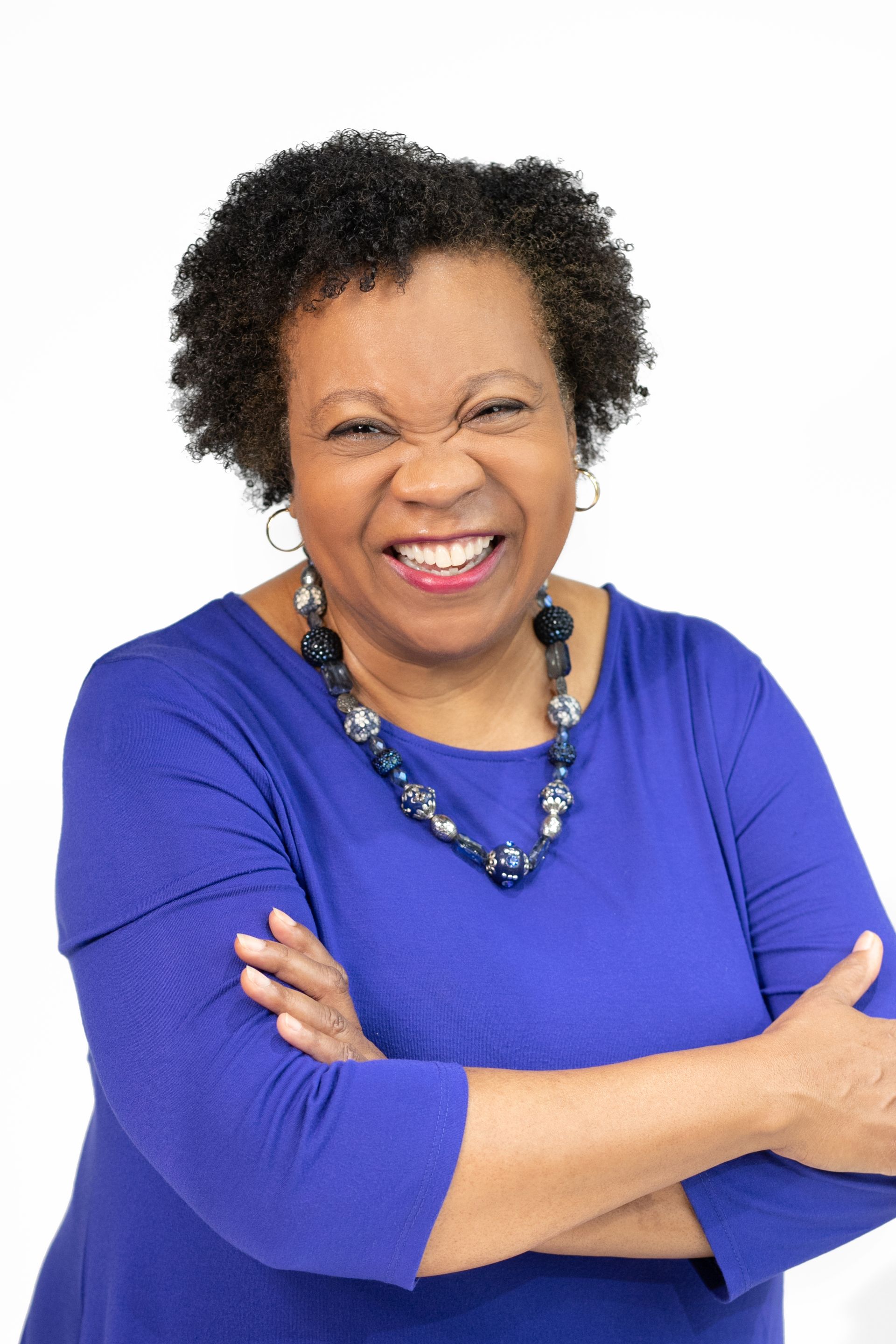 A woman in a blue shirt and necklace is smiling with her arms crossed