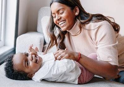 Woman smiling and tickling a laughing baby on a couch by a window