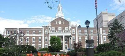 Brick civic building with white columns, central clock tower, and landscaped front entrance under a blue sky
