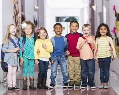 Six children standing together in a bright school hallway, smiling and holding books and backpacks.