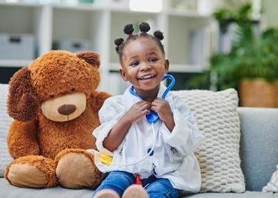 Smiling child holding a stethoscope beside a large teddy bear on a couch