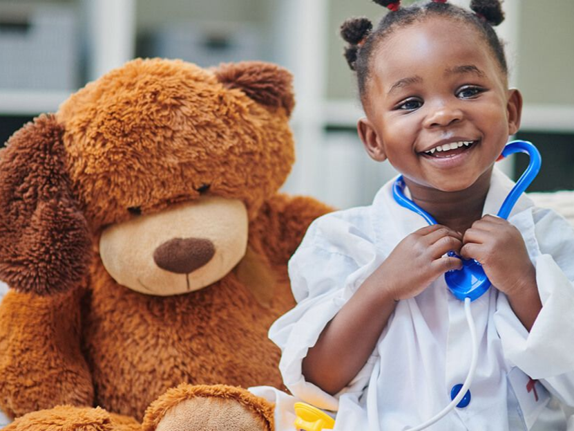 Smiling child holding a blue toy stethoscope beside a large teddy bear in a bright room