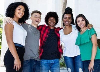 Five smiling friends standing together indoors with arms around each other.