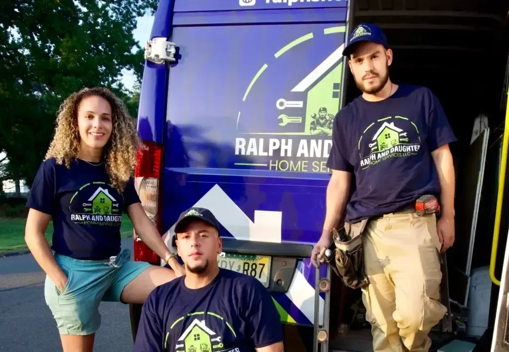 Three people posing by a blue Ralph Anthony Homes van, wearing matching company shirts.