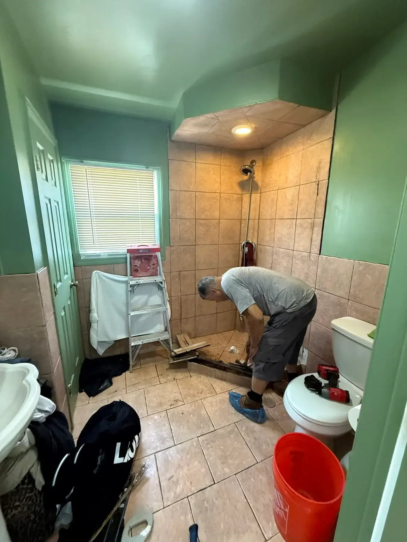 Worker tiling a small bathroom shower area, with teal walls, tan tile, and tools on the floor