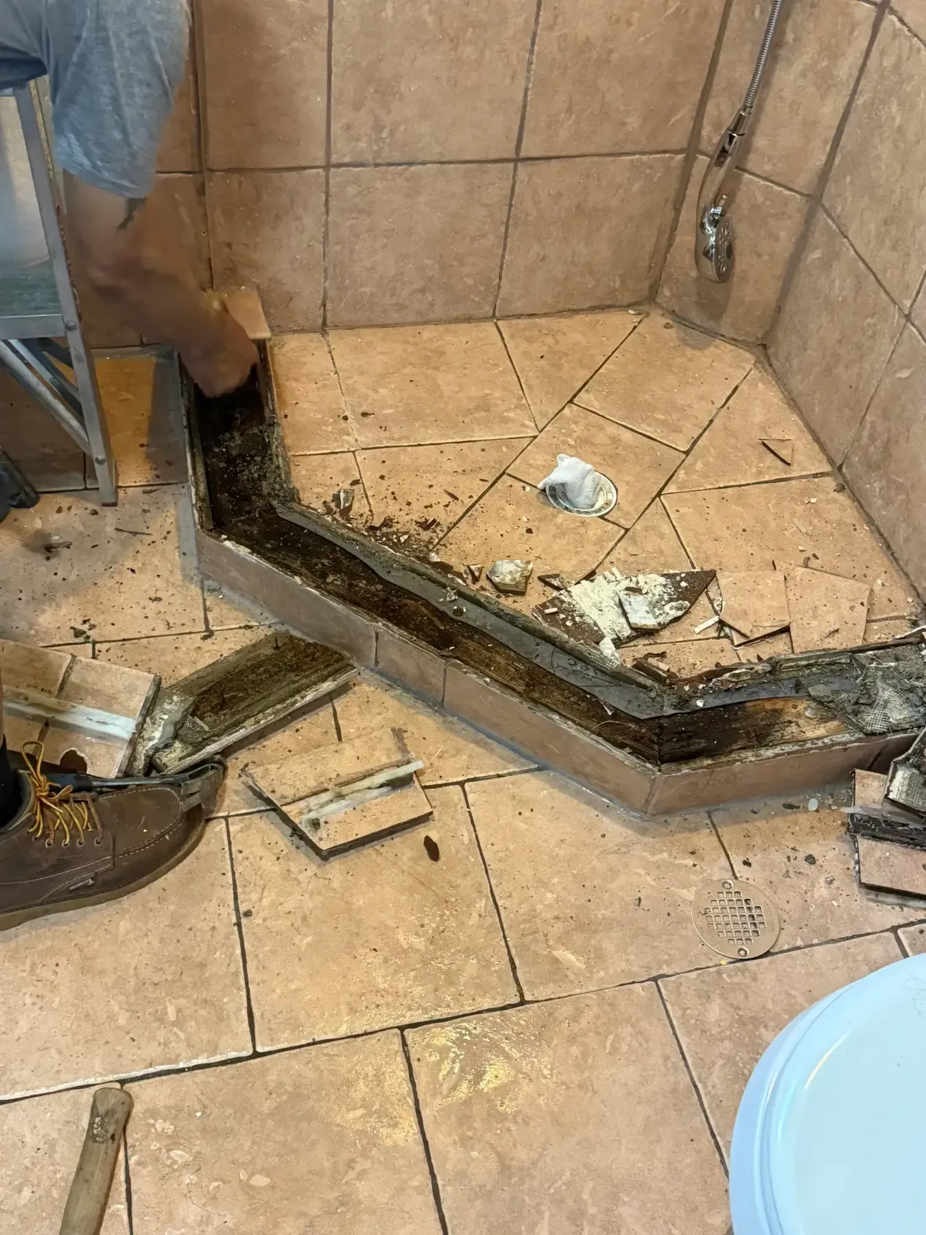 Damaged tiled shower floor with broken tiles, exposed substrate, and debris in a bathroom corner