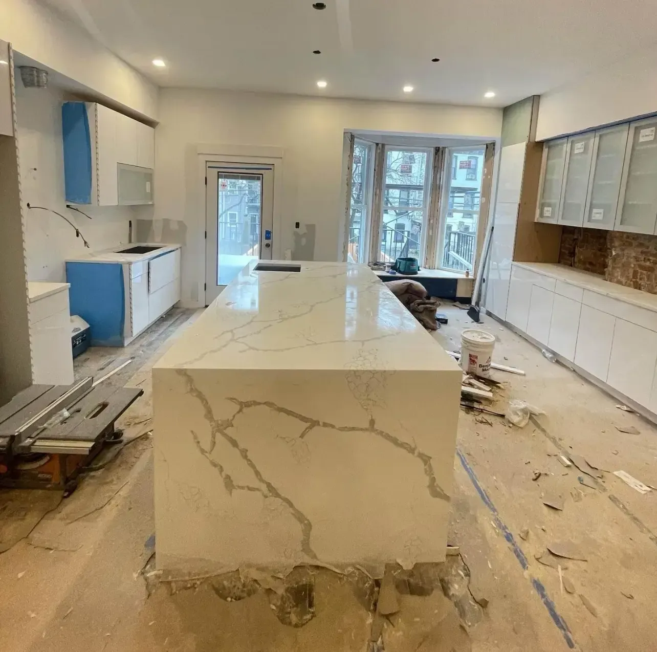 Bright kitchen under renovation with white cabinets, marble island, and construction debris on the floor
