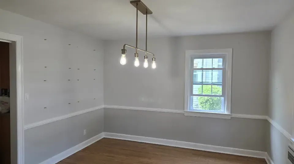 Empty dining room with white walls, wood floor, pendant light, and a small window.
