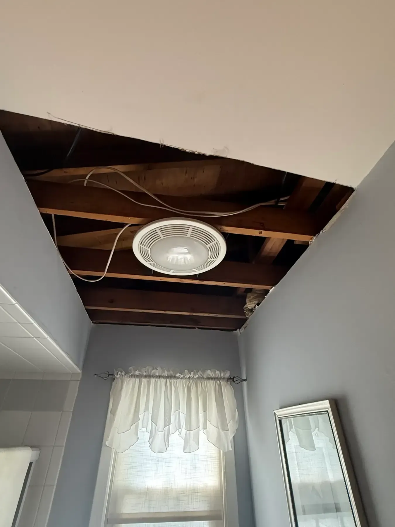 Bathroom ceiling with a large hole exposing wooden beams and a round light fixture above a curtained window